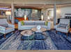 Hotel lobby with modern decor, featuring a blue-patterned rug, gray seating, two circular coffee tables, and wall art behind the sofa. A beverage station is visible on the right.