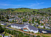 Aerial view of a suburban area with numerous houses, a large building with a pool, and green hills in the background under a clear blue sky.