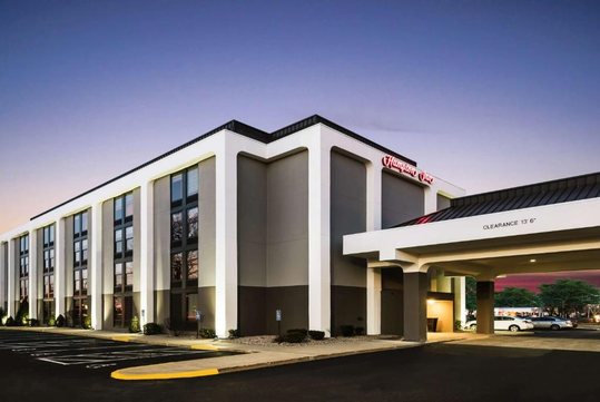 Modern hotel building with large windows, a covered entrance, and a parking lot, under a clear evening sky.