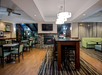 Spacious modern lounge with wooden floors, a stone fireplace, tables, chairs, and green sofas. Large windows and ceiling lights brighten the room.