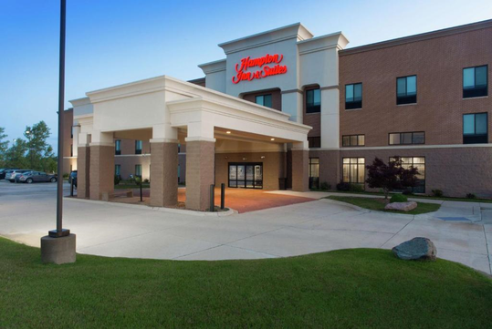 Entrance of a Hampton Inn & Suites hotel with a brick exterior, a covered driveway, and a well-maintained lawn.