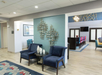 Hotel lobby with two blue armchairs and a glass table, featuring geometric wall art. Nearby, there's a dining area with colorful chairs and contemporary decor.