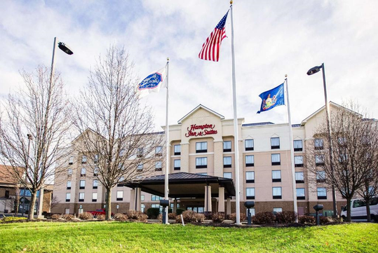 Hotel exterior with a sign reading "Hampton Inn & Suites," surrounded by three flags on flagpoles. Bare trees and a grassy area are in the foreground.