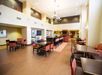 A spacious hotel dining area with tables, chairs, and a counter. The room has tall ceilings, modern decor, and warm lighting.