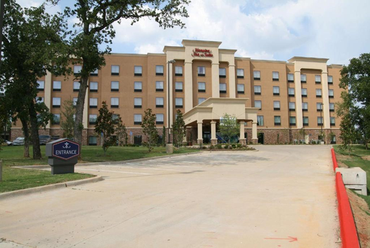 Five-story hotel building with beige exterior, surrounded by trees and a parking lot. An entrance sign is visible near the driveway.