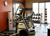 A gym room with two elliptical machines, a rack of weights, exercise balls, and a wall clock displaying the time.