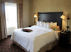 A neatly made hotel bed with white linens, four pillows, and a headboard. Two wall-mounted lamps flank the bed. A window with curtains is on the left. A tray with a remote is on the bed.
