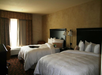 A hotel room with two double beds, a bedside table with lamps, a window with curtains, and a desk.