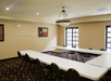 A conference room with U-shaped table setup, chairs, and a large ceiling light fixture. A flag is displayed on the wall, and natural light comes through the windows.