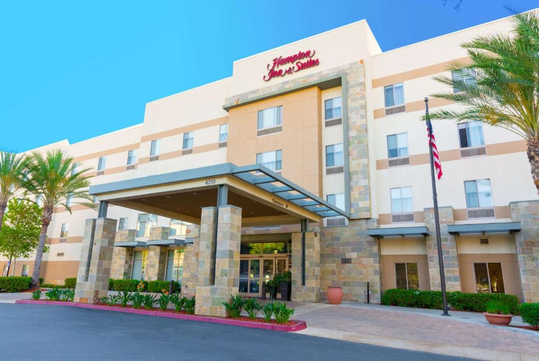 Exterior view of a Hampton Inn & Suites hotel with stone accents, palm trees, and an American flag.