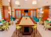 A hotel lobby with a central long table, green chairs, turquoise armchairs, and round tables. Neutral-toned walls and ceiling with pendant lights. TV and framed pictures on the wall.