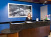 A receptionist stands behind a dark marble counter in a hotel lobby. A framed photo with the word "hello" hangs on the blue wall behind her.