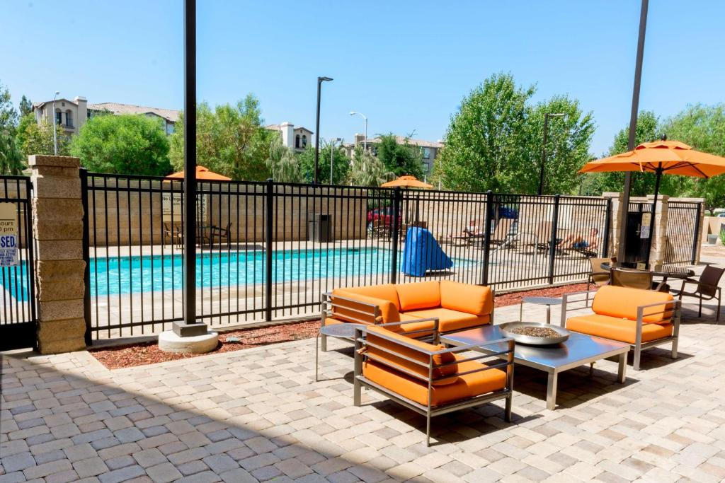 Outdoor patio with orange furniture and tables, near a fenced pool. Trees and buildings are visible in the background under a clear blue sky.