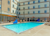 Outdoor swimming pool surrounded by a fence, with lounge chairs and tables under orange umbrellas near a multi-story building.