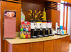 Coffee and tea station with multiple dispensers, a tea selection box, stacks of cups, and decorative vases on a wooden countertop.