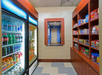 A small snack shop with a refrigerated section on the left and shelves of chips and snacks on the right, featuring a service counter in the back with a blue hallway beyond it.