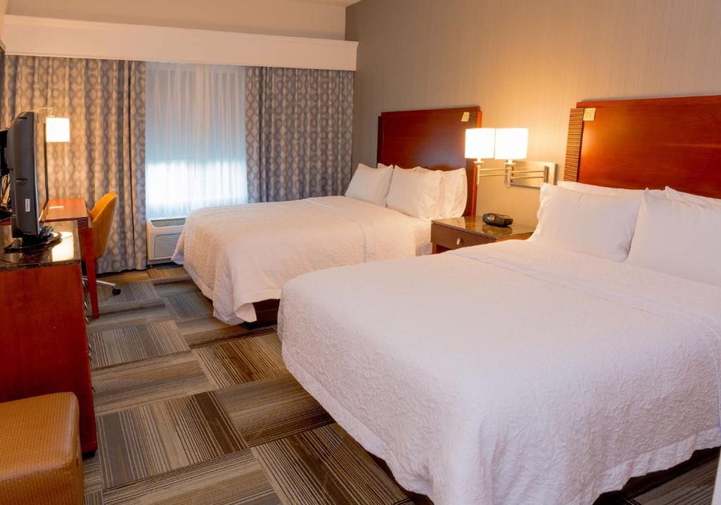 Hotel room with two double beds, wooden headboards, a TV on a stand, a desk with a chair, and patterned curtains. The room is well-lit and has a carpeted floor.
