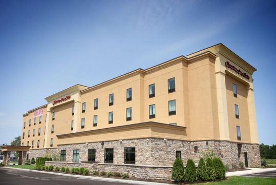 Four-story beige hotel building with stone accents and a red logo, set against a clear blue sky, surrounded by a parking lot and greenery.