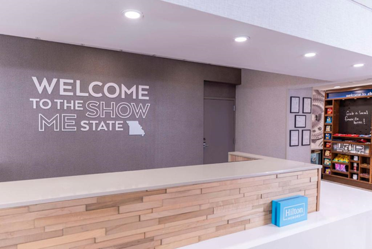 Hotel lobby with a sign that reads "Welcome to the Show Me State" and a map of Missouri. The counter has a "Hilton" sign, and a small shop is visible in the background.