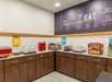 Hotel breakfast area with countertop items including a hot food station, cereal dispensers, and oatmeal packets. A chalkboard-like wall displays food-related drawings and the word "EAT.
