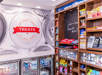 Snack shop with various treats on wooden shelves, a refrigerated section below, and a wall menu. Sign reads "TREATS.