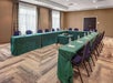 A U-shaped conference table setup with green tablecloths, water bottles, notepads, and chairs in a meeting room with carpeted floor and two windows.