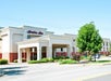 Exterior view of a Hampton Inn hotel with a covered entrance, trees, bushes, and parked cars on a sunny day.