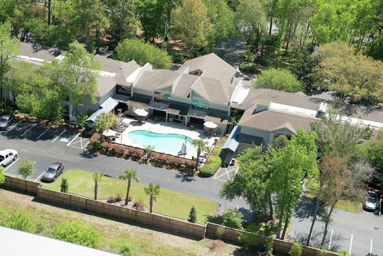 Aerial view of a residential complex with a central swimming pool, surrounded by trees. Several parked cars are visible in the adjacent parking area.