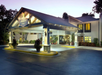 Illuminated entrance of a hotel at dusk, featuring a pitched roof and surrounding plants.
