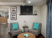 A small seating area with two gray chairs and a round wooden table, featuring teal cushions and decor. A flat-screen TV is mounted above, with framed photos on the wall.