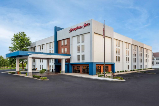 A four-story Hampton Inn hotel with a blue and white exterior, entrance canopy, American flag, landscaped grounds, and a parking lot in front.