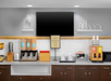 A hotel breakfast station with cereal dispensers, juice machines, a microwave, coffee maker, cups, and decorative shelves beneath a large TV screen.