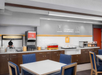 Hotel breakfast area with waffle maker, juice dispenser, covered hot food trays, toaster, microwave, and dining tables with blue chairs. White wall has a "Let's Bake Some Memories" sign.