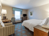 A hotel room with a king bed, desk with a lamp, accent chair, small window, and a small dresser with a TV on it.
