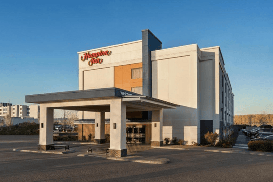 The front exterior and covered drive way and entrance of the Hampton Inn Portland Airport with the sun shining on it and clear sky over it.