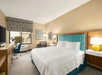 A hotel room with a white king bed with a blue head board, nightstands with lamps, an accent chair, TV on a dresser, small window, and desk and chair.