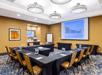 Meeting room with U-shaped table arrangement, conference chairs, wall-mounted screen displaying a bar chart, modern ceiling lights, coffee station, and framed artwork on a beige wall.