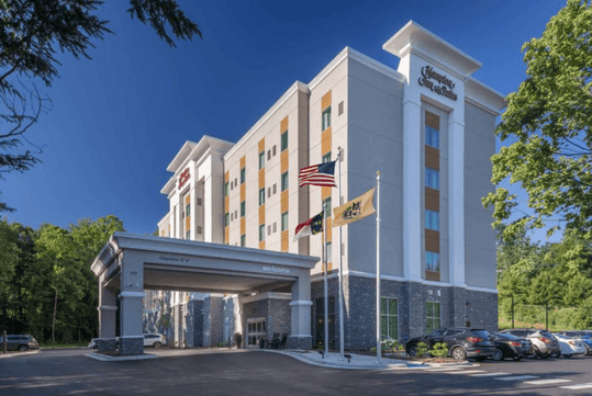 Modern hotel with five stories under a clear blue sky, featuring an entrance canopy and flags in front, surrounded by trees and parked cars.