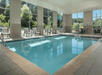 Indoor swimming pool with lounge chairs and tables around. Large windows offer a view of greenery outside.