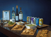 A table displays wine bottles, canned drinks, glasses, and wooden boards with snacks including edamame, pretzels, chips with dip, and fried cheese, next to a clipboard with a menu.