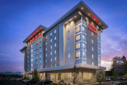 The exterior of the Hampton Inn & Suites Asheville Biltmore Area with cars parked in front of it and a purple sunset behind it.