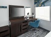 Hotel room workspace with a wooden desk, flat-screen TV, mirror, lamp, two water bottles, and a blue swivel chair. Window with geometric-patterned curtain on the right.