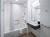 Accessible bathroom with a bathtub featuring grab bars, a showerhead, and a curtain. A sink with a mirror is visible, along with towels and a waste bin on wooden flooring.