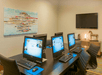 A business center with five computers on a dark brown table with office chairs in front of them, art on the left wall, and a TV on the right.