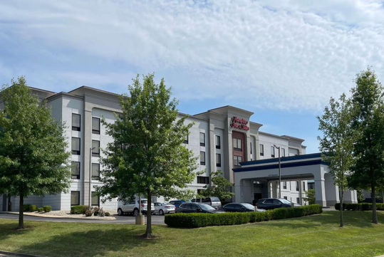 Exterior of a Hampton Inn hotel with a four-story building, parked cars, and surrounding greenery under a partly cloudy sky.