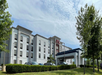 A four-story hotel building with a parking area in front, surrounded by trees and bushes under a partly cloudy sky.