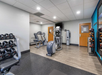 Small gym room with treadmills, an elliptical, and a stationary bike. Dumbbells are on a rack to the left. Medicine balls are on shelves in the background. Floor is a mix of wood and rubber tiles.