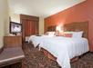 A hotel room with two white queen beds against an orange wall with a TV sitting on a dresser and a bench across from them.