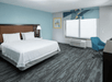 A modern hotel room with light blue and gray decorations, a white king bed with lamps in the headboard, a large window, and desk and chair.