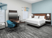 A modern hotel room with a light blue accent wall, a white king bed, two small windows, and a desk with a bright blue office chair.